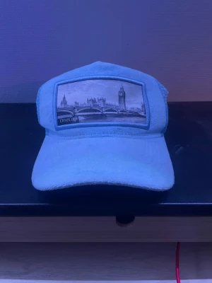 Ljusblå keps med Londonmotiv - Snygg ljusblå keps från One Cap med mesh på sidorna och broderad patch framtill med Londonmotiv, inklusive Big Ben och Tower Bridge. Kepsen har böjd skärm och en liten metallbricka på sidan. Perfekt för dig som gillar streetstyle och unika accessoarer.