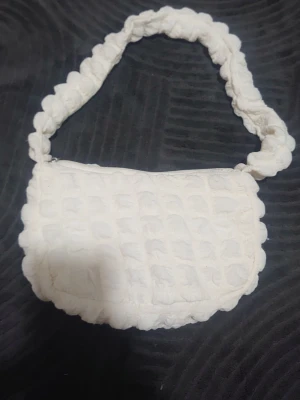 Vit quiltad axelväska med bubblig rem - Supersöt axelväska i vit quiltad textil med bubblig struktur och matchande vadderad axelrem. Väskan har dragkedja upptill och ett rymligt innerfack. Perfekt för dig som gillar unika accessoarer med mjuk känsla.