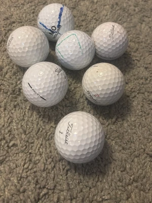 7 vita Titleist golfbollar - Säljer sju stycken vita golfbollar från Titleist, flera med tryck som Pro V1 och olika nummer. Bollarna har klassisk dimple-yta och några har märkningar med penna. Perfekt för dig som vill fylla på golfbagen.