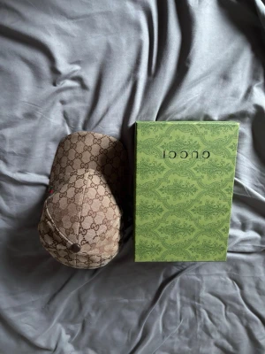 Gucci keps med GG-mönster - Säljer en snygg beige keps från Gucci med klassiskt GG-monogram över hela kepsen. Kepsen har gröna och röda detaljer på sidan och justerbar rem bak. Materialet är canvas med läderdetaljer. Perfekt för dig som vill ha en lyxig accessoar med ikonisk design.