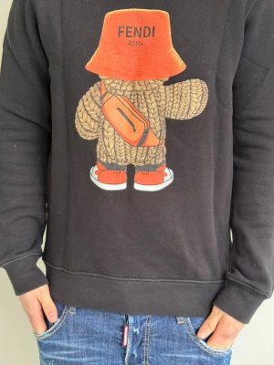 Svart Fendi hoodie med grafiskt tryck - Svart hoodie från Fendi med ett stort grafiskt tryck framtill av en figur med orange hatt, väska och skor. Tröjan har rund halsringning, långa ärmar och ribbade muddar. Materialet är mjukt och bekvämt, perfekt för en avslappnad streetwear-look. Han på bild är 175cm och väger 74kg