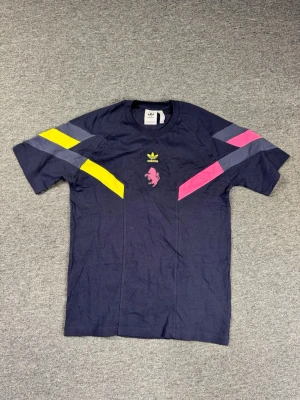 Adidas Juventus mörkblå t-shirt - Mörkblå t-shirt från Adidas x Juventus med färgglada detaljer i gult, rosa och grått på axlarna. Bröstet har Adidas-logga och Juventus-lejon i rosa. Tillverkad i mjukt bomullsmaterial och har en sportig, avslappnad passform.