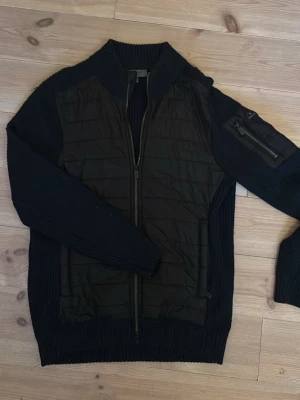 Svart cardiganjacka från Snoot - Stilren svart cardiganjacka från Snoot med quiltad front och stickade ärmar. Jackan har dragkedja framtill, ribbade muddar och en praktisk ficka med dragkedja på ärmen. Perfekt för lager-på-lager och snygg till jeans eller chinos.