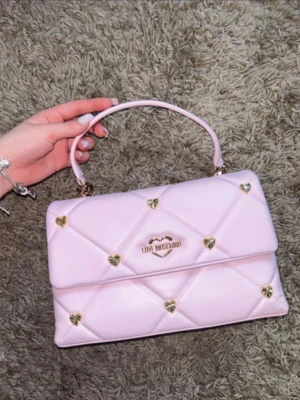 Rosa quiltad handväska Love Moschino - Superfin ljusrosa quiltad handväska från Love Moschino med guldfärgade hjärtdetaljer och logga framtill. Väskan har ett topphandtag och är tillverkad i ett glansigt konstlädermaterial. Perfekt för dig som gillar söta och trendiga accessoarer. Ord. Pris 2300kr