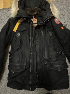 Svart Parajumpers parkas med pälskrage - Svart parkajacka från Parajumpers med pälskantad huva, flera fickor och ikonisk gul rem vid kragen. Jackan har quiltat foder och detaljer i skinn samt dragkedja och knappar framtill. Perfekt för kalla dagar.