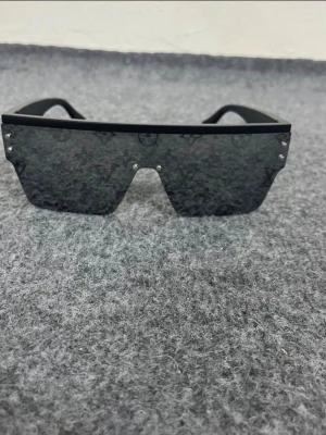  Louis Vuitton solglasögon - Säljer ett par svarta shield-solglasögon från Louis Vuitton med monogrammönster på glaset. De har en futuristisk, kantig form och breda skalmar. Glasen är mörka och bågarna är i blank plast. Perfekta för dig som vill sticka ut med en exklusiv accessoar.