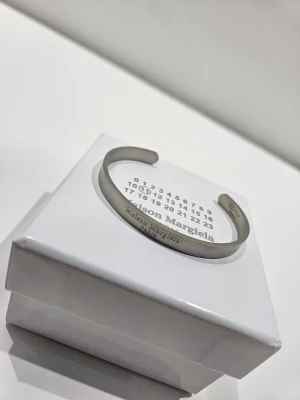 Maison Margiela silverfärgat armband - Stilrent armband i silverfärgad metall från Maison Margiela. Öppen design med ingraverad logotyp på utsidan. Kommer i original ask med märkets ikoniska siffror. Perfekt accessoar för en minimalistisk och modern look.