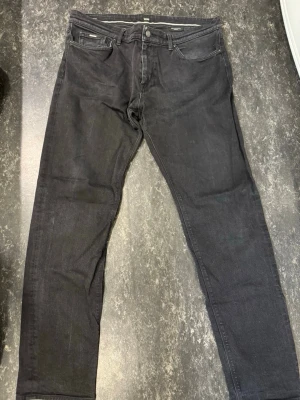 Svarta slim fit jeans från boss - Snygga svarta slim fit jeans med klassisk femficksdesign och dragkedjegylf. Jeansen har raka ben och diskreta sömmar, perfekta till en avslappnad stil. Materialet är stretchigt för extra komfort. Liten skada på benet (se bild)