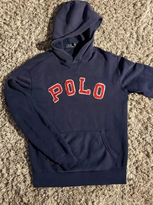 Marinblå hoodie från Polo Ralph Lauren🙌🤩 - Snygg marinblå hoodie från Polo Ralph Lauren med stor röd POLO-text i frotté på bröstet. Klassisk känguruficka framtill och huva. Perfekt för en chill och sportig look. Tillverkad i mjuk bomullsblandning för extra komfort.🙌🤩
