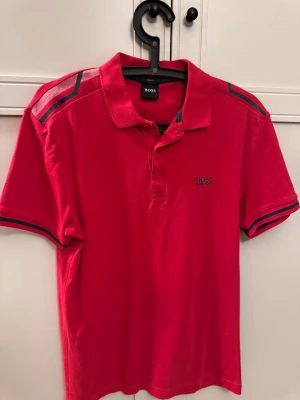 Röd pikétröja Hugo Boss - Snygg röd piké från BOSS med klassisk krage och knappar framtill. Tröjan har mörkblå detaljer på axlar och ärmslut samt diskret BOSS-logga på bröstet. Perfekt för en stilren och sportig look.