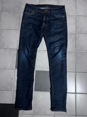 Mörkblå jeans från Nudie Jeans - Snygga mörkblå jeans från Nudie Jeans med klassisk femficksdesign och guldfärgade sömmar. Jeansen har en smal passform och är tillverkade i denim med lätt slitning på låren. Bakfickan har Nudie Jeans karakteristiska brodyr och läderpatch i guld. Längd 31 och midja 34. Hör av er vid funderingar eller frågor. Fraktar samma dag om det köps innan klockan 2 på eftermiddagen💨