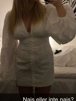 Vit kort klänning från Adoore - Supersöt vit kort klänning från Adoore med v-ringning och långa puffärmar. Klänningen har diskret blommigt mönster i tyget och knappar framtill. Perfekt för student eller skolavslutning