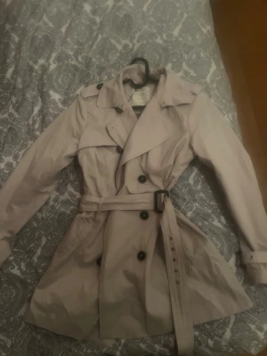 Vintage Beige trenchcoat med bälte - Stilren beige trenchcoat med klassisk dubbelknäppning, bred krage och bälte i midjan. Jackan har svarta knappar, axelklaffar och fickor på sidorna. Köpt på secondhand i Köpenhamn för 399kr ( Från street one )