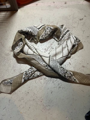 Beige och vit mönstrad halsduk - Snygg halsduk i beige och vitt med coolt grafiskt mönster av rutor, prickar och linjer. Tunt och lätt material som känns mjukt mot huden. Perfekt att knyta runt halsen eller använda som accessoar för att lyfta din outfit.