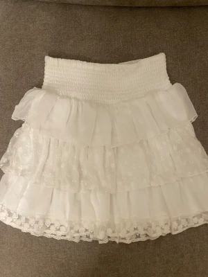 Vit volangkjol med spetsdetaljer - Supersöt vit kortkjol med flera lager volanger och broderade blommor. Kjolen har bred smock i midjan för skön passform och avslutas med spetskant längst ner. Perfekt för dig som gillar romantisk och feminin stil. st 148-152