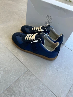 Maison Margiela sneakers blå - Maison Margiela sneakers i mörkblå mocka och skinn med vita snören och beige innersula. Klassisk siluett med gummisula och diskret branding på plösen. Snygg retrostil som passar perfekt till streetwear eller casual outfits.