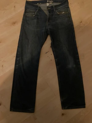 Mörkblå jeans från Jack & Jones - Snygga mörkblå jeans från Jack & Jones med klassisk femficksdesign och bruna detaljer på knappar och läderpatch. Jeansen har rak passform och är tillverkade i slitstarkt denim med kontrastsömmar. Perfekt för dig som gillar enkel och stilren look.
