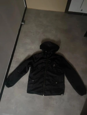 Trapstar whindbraker - Svart vindjacka med huva från True Saker. Jackan har en snygg silverfärgad dragkedja och logotyp på bröstet. På ryggen finns broderad text. Materialet är lätt och syntetiskt, perfekt för blåsiga dagar. Långärmad och med normal passform.