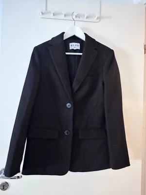 Svart kavaj från NA-KD - Stilren svart kavaj från NA-KD med klassisk krage och två knappar framtill. Kavajen har raka ärmar och två fickor med lock, perfekt för en snygg och enkel look. Materialet känns mjukt och har en matt finish.