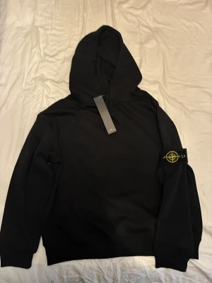 Svart hoodie från Stone Island - Svart hoodie från Stone Island med klassisk patch på vänster ärm. Tröjan har huva, ribbade muddar och en relaxed passform. Materialet är mjuk bomull och designen är stilren utan mönster. Perfekt för dig som gillar streetwear och vill ha något snyggt och bekvämt.