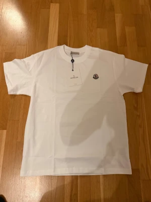 Vit Moncler t-shirt med logga - Snygg vit t-shirt från Moncler i storlek L. Klassisk design med rund hals och korta ärmar, tillverkad i mjuk bomull. På bröstet sitter Monclers ikoniska logotyp som ger en exklusiv touch. Perfekt för dig som gillar stilrena och lyxiga plagg.