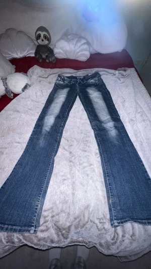 Blå bootcut jeans från New Age - Snygga blå jeans från New Age med bootcut passform och slitna detaljer framtill. Jeansen har låg midja, dragkedjor på fickorna och coola kontrastsömmar. Baksidan har broderade fickor med vita detaljer och en patch med blommotiv. Säljer pga förstora❤️                                      Midjemått: ca 34cm men stretch                          Innerbensländ: 80-81 cm  