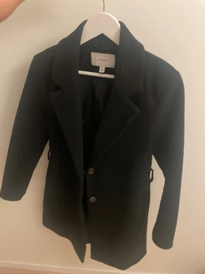 Svart kappa från VILA, storlek 36 - Snygg svart kappa från VILA i klassisk modell med två knappar framtill och krage. Jackan har lång ärm och stilren design som passar till många olika outfits. Materialet är mjukt och känns skönt mot kroppen. Perfekt för dig som gillar enkel och tidlös stil.