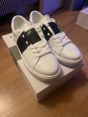 Valentino Garavani vita sneakers - Snygga vita sneakers från Valentino Garavani med bred svart rem över ovansidan och coola nitar på hälen. Skorna har rund tå, platt sula och klassisk snörning. Tillverkade i slätt skinn med vita och svarta detaljer som ger en stilren och modern vibe. Sprillans nya bara testade, kan pruta ned lite!