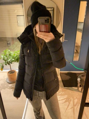 pufferjacka från Polo Ralph Lauren - Mörkblå pufferjacka från Polo Ralph Lauren med klassisk logga på bröstet. Jackan har hög krage, huva och dragkedja framtill. Fylld med dun och har två sidofickor med knappstängning. Perfekt för kalla dagar och har en snygg, figurnära passform.