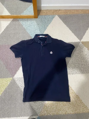 Mörkblå Moncler pikétröja i bomull - Snygg mörkblå pikétröja från Moncler med klassisk krage och korta ärmar. Tröjan har knappar framtill och Moncler-logga broderad på bröstet. Röd och vit randig detalj på insidan av kragen och vid knäppningen. Perfekt för dig som gillar stilrena märkesplagg.