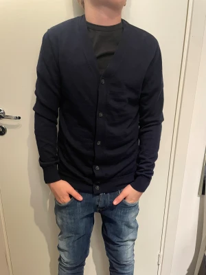 Mörkblå kofta från These Glory Days - Snygg mörkblå kofta från These Glory Days med klassisk v-ringning och knappar framtill. Koftan är långärmad och har ribbade muddar vid ärmslut och nederkant. Perfekt för lager-på-lager och skandinavisk stil.