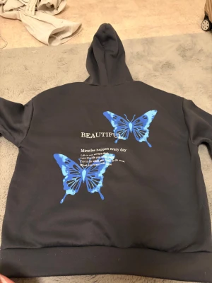 Mörkgrå hoodie med blå fjärilar - Mörkgrå hoodie med huva och tryck på ryggen. Två stora blå fjärilar och texten 'BEAUTIFUL' samt inspirerande citat. 