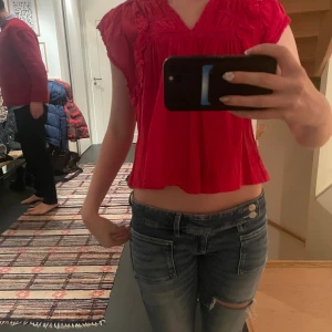 Röd blus med volanger från Neo Noir - Säljer en röd blus från Neo Noir med v-ringning, volangdetaljer och smockade partier. Blusen är kort och har ärmlös design med romantiska spetsdetaljer och luftig känsla. Perfekt för dig som gillar färgstarka och feminina plagg.