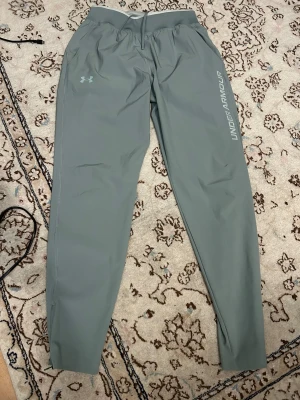 Grå träningsbyxor från Under Armour - Snygga gljusgröna träningsbyxor från Under Armour med logga på benet och diskret tryck på låret. Byxorna har elastisk midja med snörning och är tillverkade i lätt syntetmaterial som andas. Perfekta för gymmet eller löprundan.