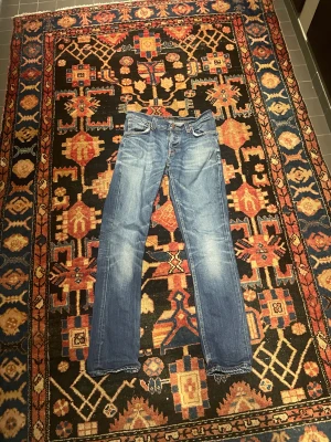 Nudie jeans blå straight fit/ slim fit - Snygga Nudie jeans i klassisk blå tvätt med lätt slitningar och raka ben. Jeansen har fem fickor, detaljerade sömmar och läderpatch bak i midjan. Passformen är straight och materialet är slitstark jeansbomull.