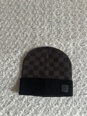  rutig mössa i svart - Snygg mössa med klassiskt rutigt mönster i mörkgrått och svart. Mössan har en ribbad svart kant och en diskret plakett framtill. Perfekt accessoar för dig som gillar exklusiv streetstyle och vill sticka ut med designerplagg.