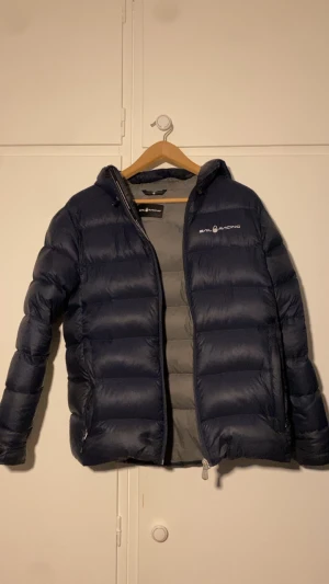 Mörkblå dunjacka från Sail Racing - Snygg mörkblå dunjacka från Sail Racing med huva och logga på bröstet. Jackan har quiltad design, dragkedja framtill och två sidofickor med dragkedja. Perfekt för kalla dagar och har en clean look som funkar till de flesta stilar. 