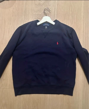 Mörkblå sweatshirt från Polo Ralph Lauren - Snygg mörkblå sweatshirt från Polo Ralph Lauren . Tröjan har rund hals, långa ärmar och ribbade muddar vid ärmslut och nederkant. Perfekt för chill dagar och enkel att matcha med jeans eller joggers.