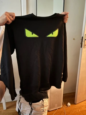 Svart Fendi tröja med gröna ögon - Cool svart tröja från Fendi i storlek XXL med unika gröna ögon på bröstet. Tröjan har lång ärm och är gjord i mjuk bomull, perfekt för dig som vill sticka ut med en statement piece. Passformen är normal och designen är riktigt edgy.