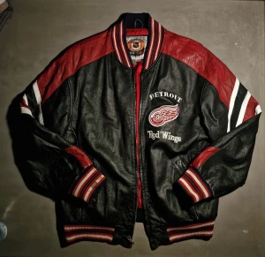 Detroit Red Wings vintage läderjacka - Säljer en otrolig svart och röd jacka i skinn med Detroit Red Wings broderat på både fram- och baksidan. Jackan har vita detaljer, ribbade muddar och dragkedja. Snygga stripes på ärmarna och klassisk sportig vibe. Dragkedjan strular en del och det finns tecken på använding! 