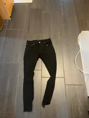 Svarta skinny jeans från Nudie Jeans - Säljer ett par svarta skinny jeans från Nudie Jeans. De har klassisk femficksdesign, smal passform och är tillverkade i stretchig bomullsmix för extra komfort. Snygga detaljer med nitar och svarta sömmar. Perfekta till sneakers eller boots. W32/l30