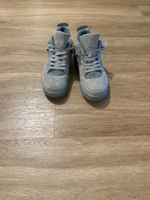 Nike Air Jordan 4 x Kaws grå sneakers - Nike Air Jordan 4 x Kaws sneakers i grå mocka med unika broderade detaljer och snörning. Skorna har platt sula, rund tå och coolt Kaws-mönster över hela ovandelen. Perfekt för dig som vill sticka ut med limited edition sneakers.