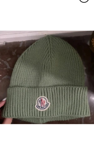 Grön stickad mössa - Snygg grön ribbstickad mössa med uppvikt kant och klassisk logopatch framtill.Passar till både jacka och hoodie. Aldrig använd