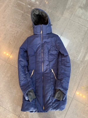 Marinblå lång dunjacka Equestrian XS - Lång marinblå dunjacka från Equestrian Stockholm med huva, dragkedja framtill och två snedställda fickor med dragkedja. Jackan har quiltad design, svarta muddar med tumhål och broderad logga på ryggen. Perfekt för kalla dagar.  (Knappt använd, ny pris 3500)