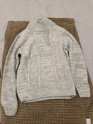 Grå stickad tröja LCW Casual - Säljer en grå stickad tröja från LCW Casual med snyggt kabelmönster framtill och hög krage med snörning. Tröjan har långärmade ärmar och ribbade muddar vid ärmslut och nederkant. Perfekt för dig som gillar mysiga och stilrena plagg.