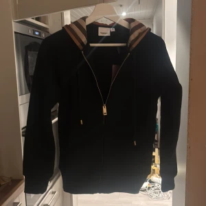 Svart Burberry hoodie - Svart hoodie från Burberry med klassisk rutig huva i beige, brunt och rött. Tröjan har dragkedja framtill, guldfärgade detaljer och snörning vid huvan. Långärmad och i mjukt bomullsmaterial, perfekt för en chill och stilren look.
