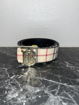 bälte med rutigt mönster - Säljer ett bälte med klassiskt rutigt mönster i beige, svart och rött. Bältet har en stor guldfärgad metallspänne med en ikonisk 'B'-logo. Ena sidan är svart med präglat mönster och den andra är beige med rutor. Perfekt för att lyfta din outfit.