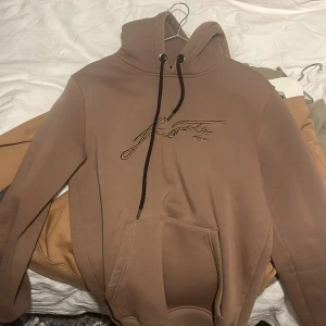 Beige hoodie från Lacoste med broderi pris kan diskuteras - Snygg beige hoodie från Lacoste med broderad logga i samma ton på bröstet. Tröjan har huva med svarta snören och en stor magficka. Mjuk insida i fleece och klassisk passform, perfekt för chill dagar. Stilren och enkel att matcha med jeans eller joggers.