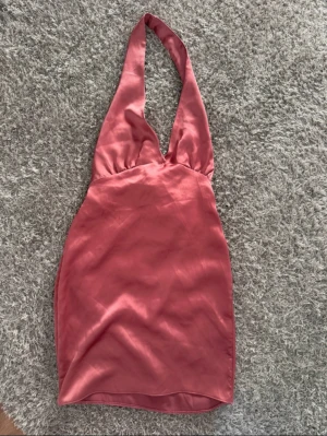 Rosa halterneck klänning PrettyLittleThing - Snygg kort halterneck klänning i skimrande rosa satin från PrettyLittleThing. Klänningen har djup v-ringning, breda axelband och knytband i midjan för en extra detalj. Perfekt för dig som vill sticka ut med en trendig och elegant look.