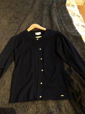 Marinblå kofta från Holly & Whyte - Snygg marinblå kofta från Holly & Whyte by Lindex i storlek S. Koftan är stickad med ribbade detaljer, har långärmade puffärmar och guldiga knappar framtill. Kolla sista bilden ett litet hål som går lätta att fixa men den syns även inte!!